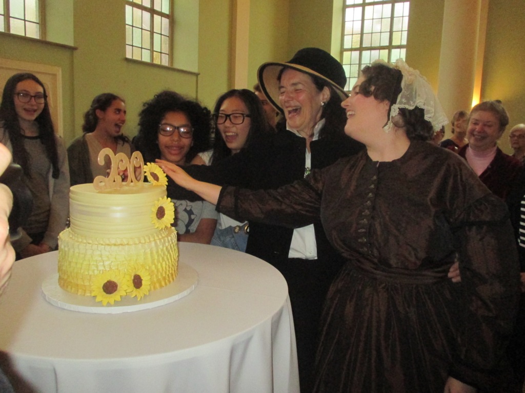 JENKINS, (CENTER) CLAD IN period dress, huddles around a birthday cake for her great-great grandmother, Elizabeth Cady Stanton, alongside guests. Photo by Vivian Carter